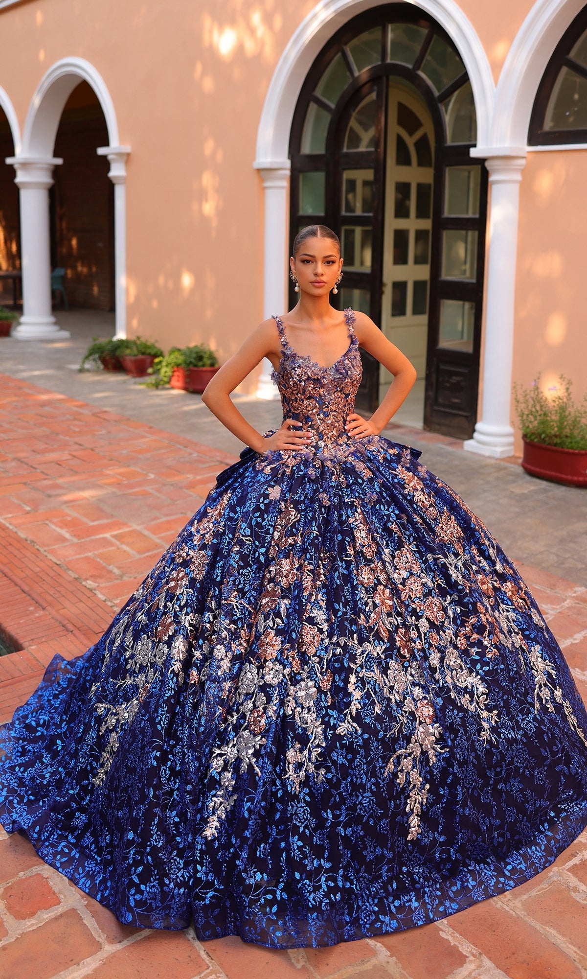 Outdoors, a person wears the Quinceanera Dress 54322 By Amarra?a voluminous blue ball gown with floral embroidery and intricate silver and gold accents.